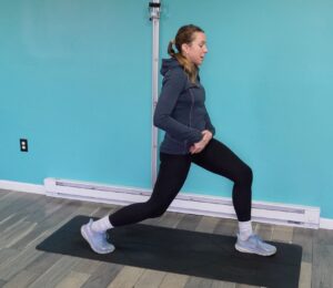 Dr. Chloe in a split stance with her right leg back, left foot in front with her back knee slightly bent.  She is pointing to her right hip as she tucks her pelvis to stretch that right side.  